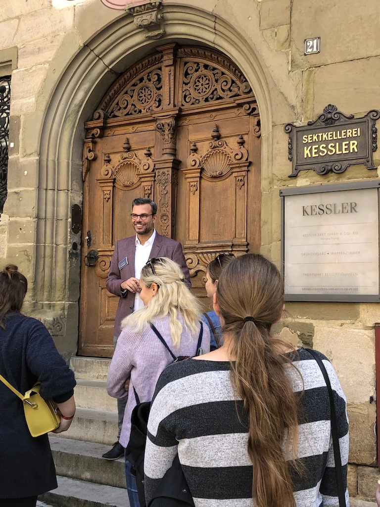 Andreas Schnug lächelt bei der Begrüßung einer Gästegruppe vor der großen Holztür der Sektkellerei Kessler